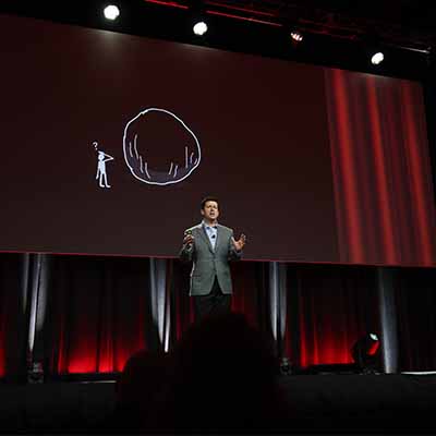 Man on stage speaking to a crowd with a graphic of a boulder and man with question mark above his head on the screen