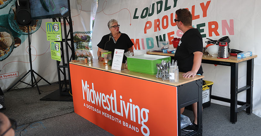 A woman and man talking with a sign behind them that reads 'Loudly Proudly Midwestern".. They are at a table that has food and culinary items on it.